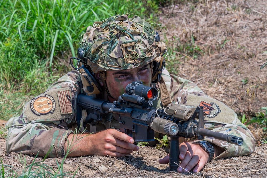 An airman lying in a field looks through the scope of a weapon.
