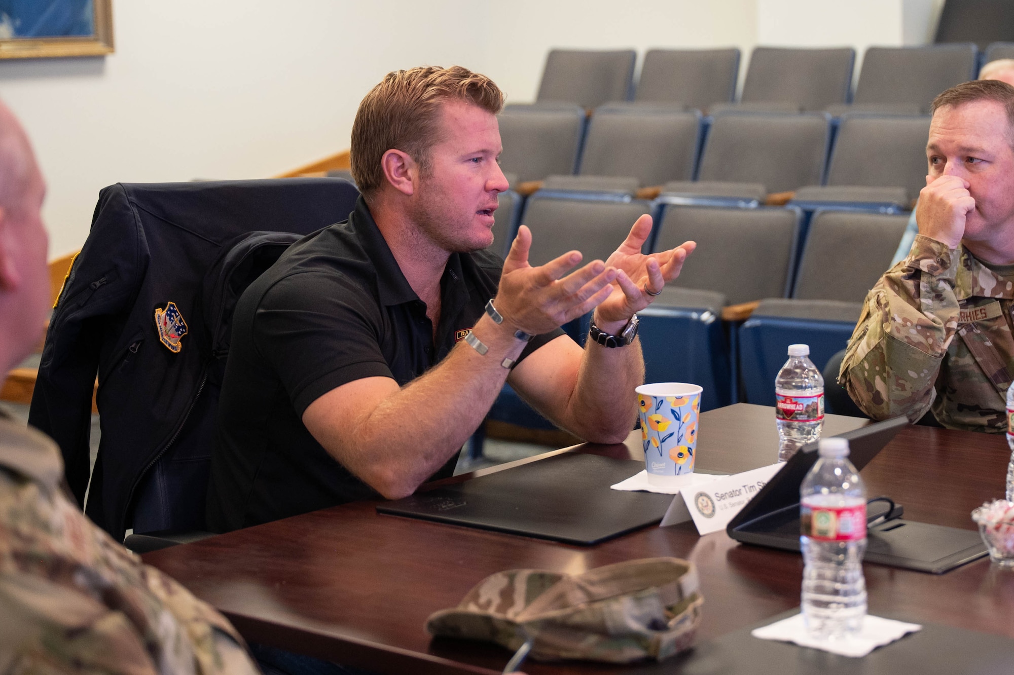 A man in civilian attire speaks to a table of men in uniform.