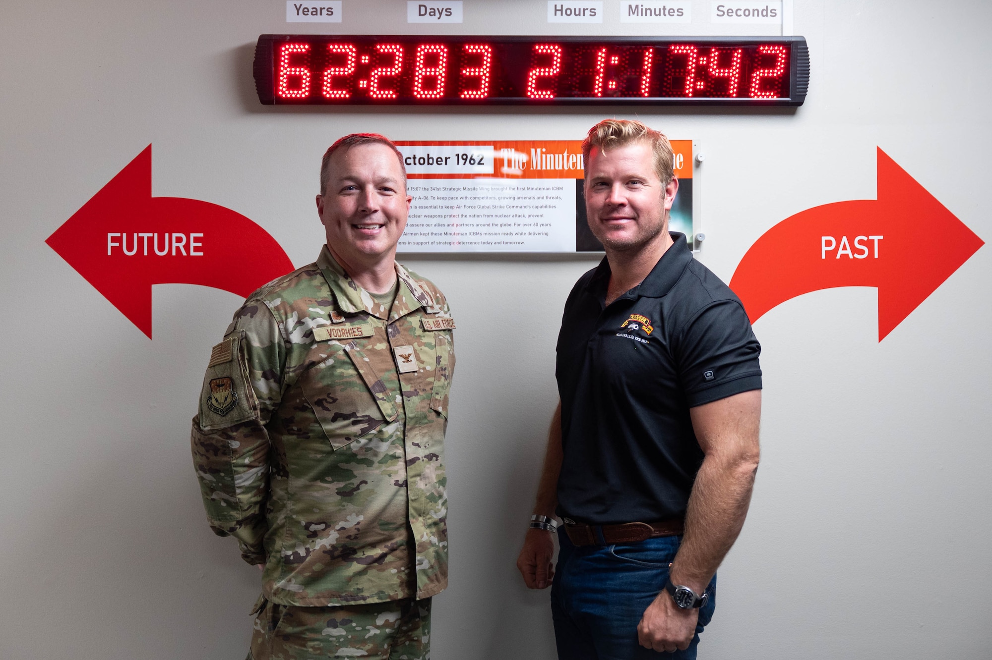 A man in uniform stands next to a man in civilian attire.