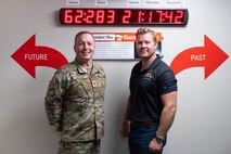 A man in uniform stands next to a man in civilian attire.