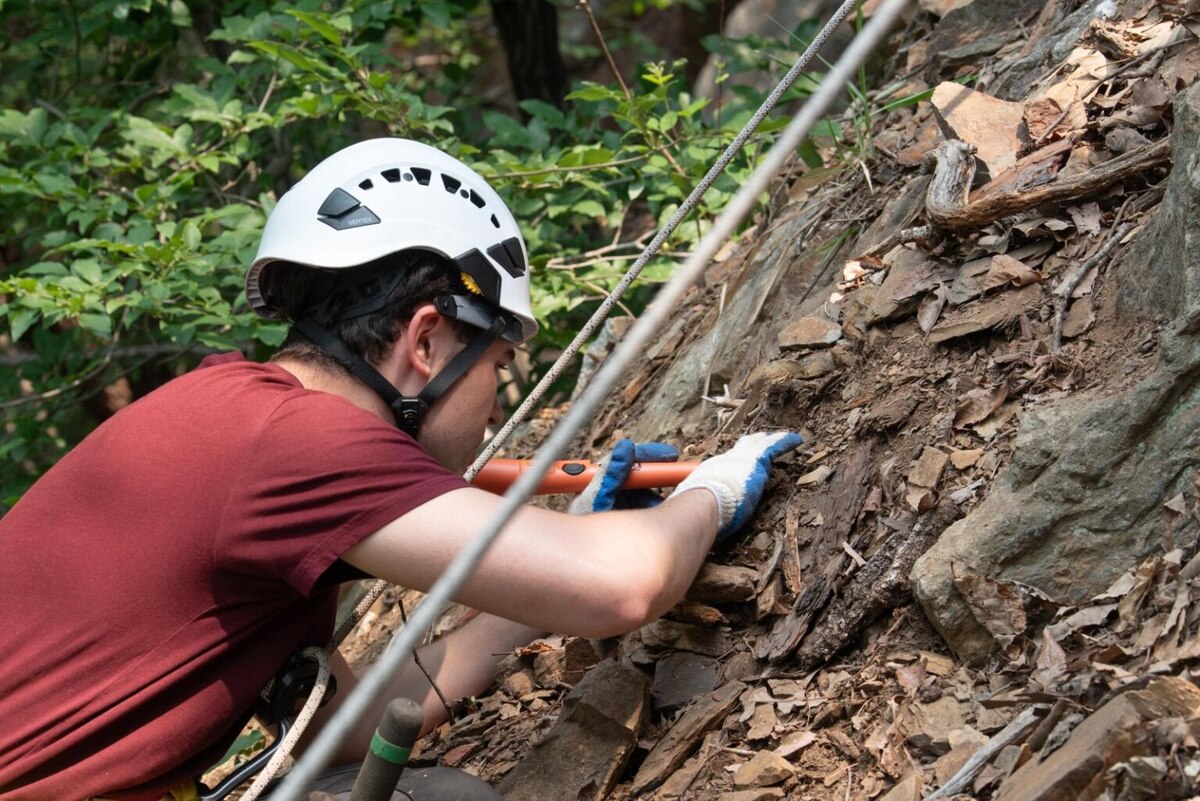 A man wearing a T-shirt and a helmet uses a tool to dig in dirt and gravel. He is attached to cables.