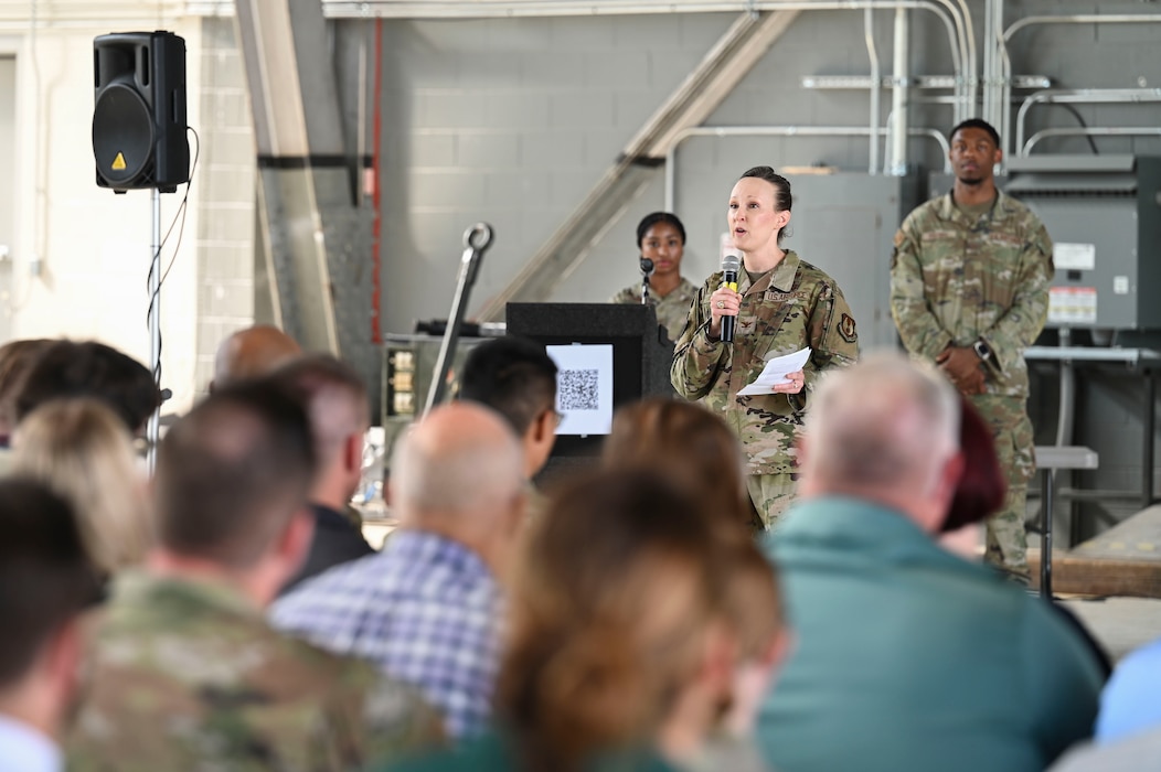 A woman in a camouflage uniform speaks into a microphone to a crowd of people wearing both military uniforms and civilian clothes.