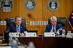 Two men sit in chairs at the head of a table in a conference room.