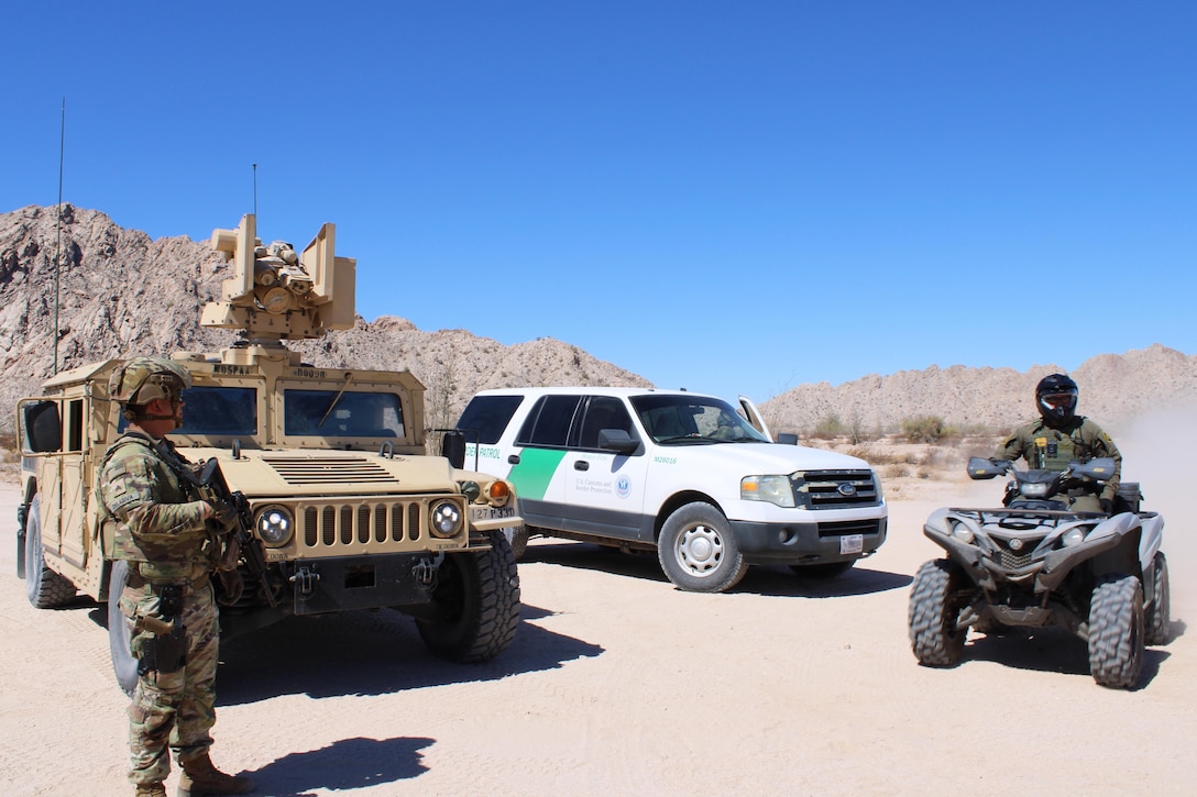 A soldier standing near a tactical vehicle talks to a border agent on a four-wheeler near a sport-utility vehicle in desert-like conditions on a sunny day.