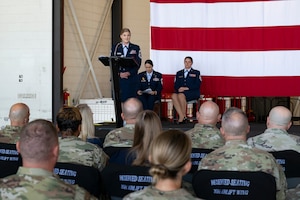 A woman in military service dress standing at a podium on stage with large American flag in the background speaking to a military audience.