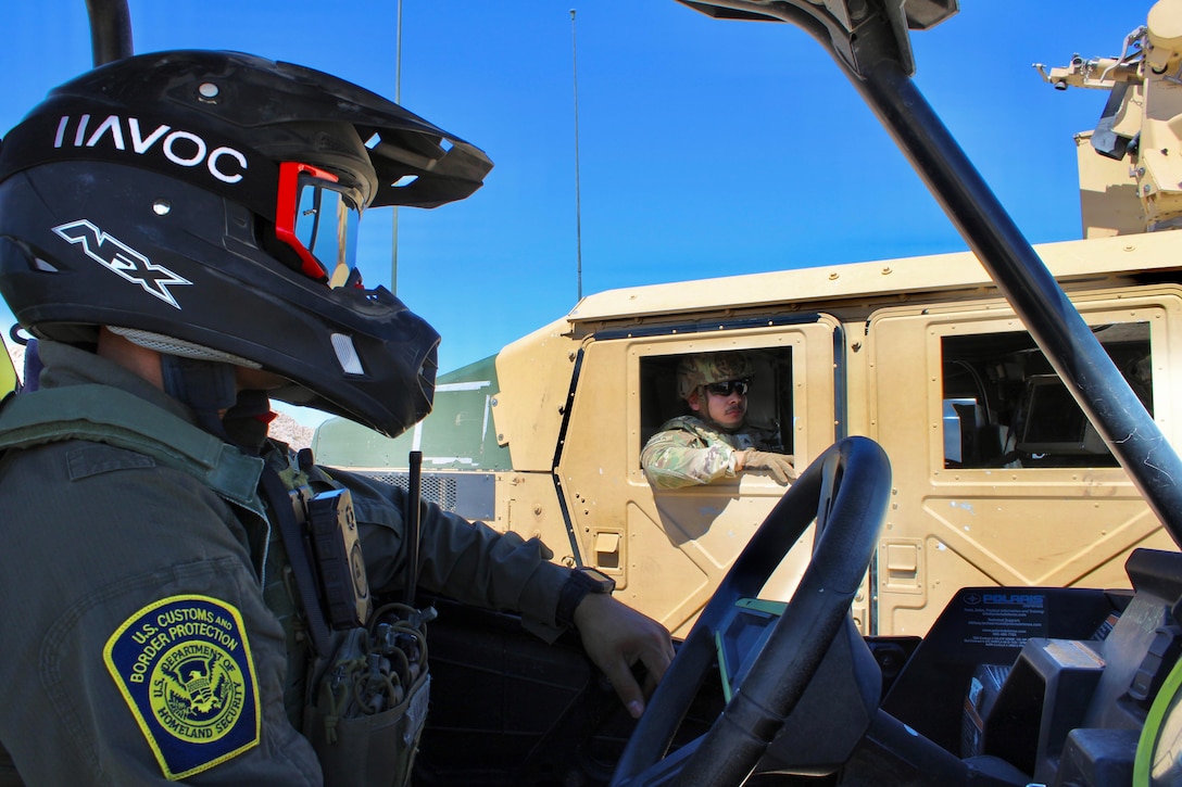 A border agent wearing a helmet and goggles inside a small vehicle talks to a soldier sitting in the back of a tactical vehicle on a sunny day.