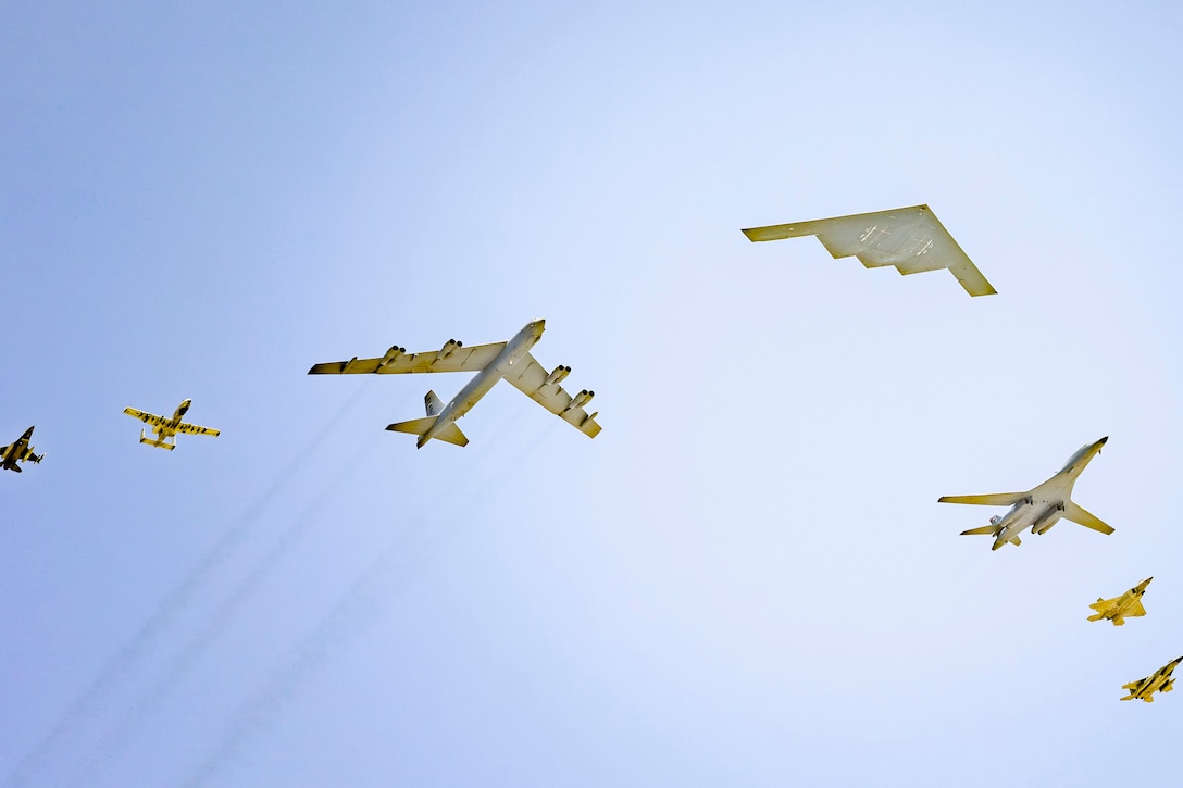 Seven different types of military aircraft fly overhead on a clear blue sky.