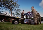 Six people wearing camouflage uniforms stand in a field holding an American flag above a casket wrapped in plastic. A truck is on their left, and a tree is behind them.