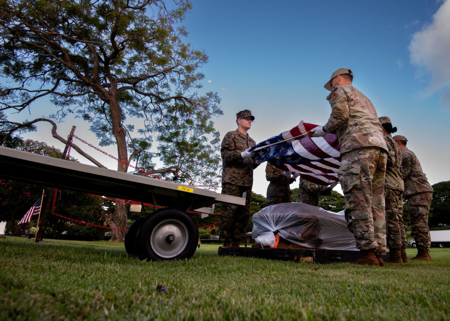 Six people wearing camouflage uniforms stand in a field holding an American flag above a casket wrapped in plastic. A truck is on their left, and a tree is behind them.