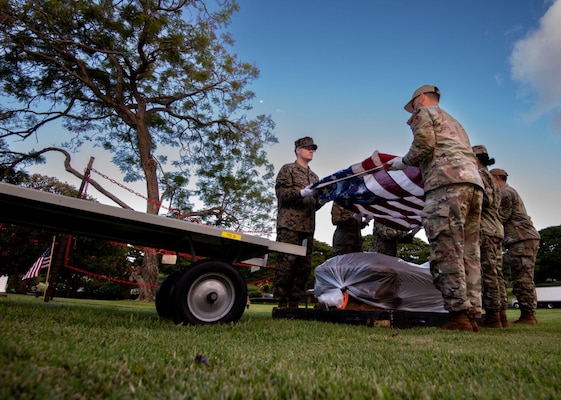 Six people wearing camouflage uniforms stand in a field holding an American flag above a casket wrapped in plastic. A truck is on their left, and a tree is behind them.