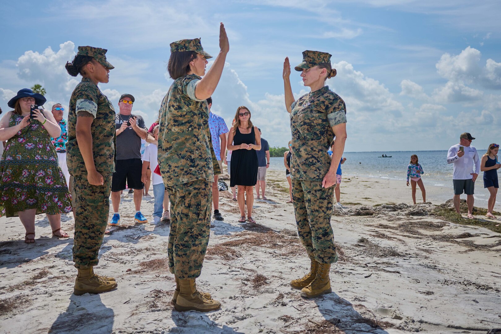 Two Marines raise their right hands on the sand while family members watch in front of the beach.