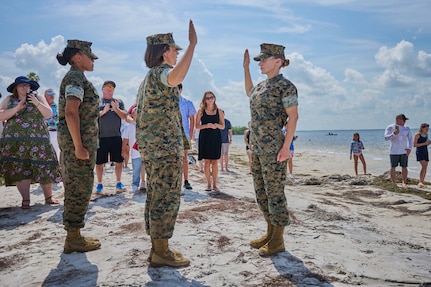 Two Marines raise their right hands on the sand while family members watch in front of the beach.