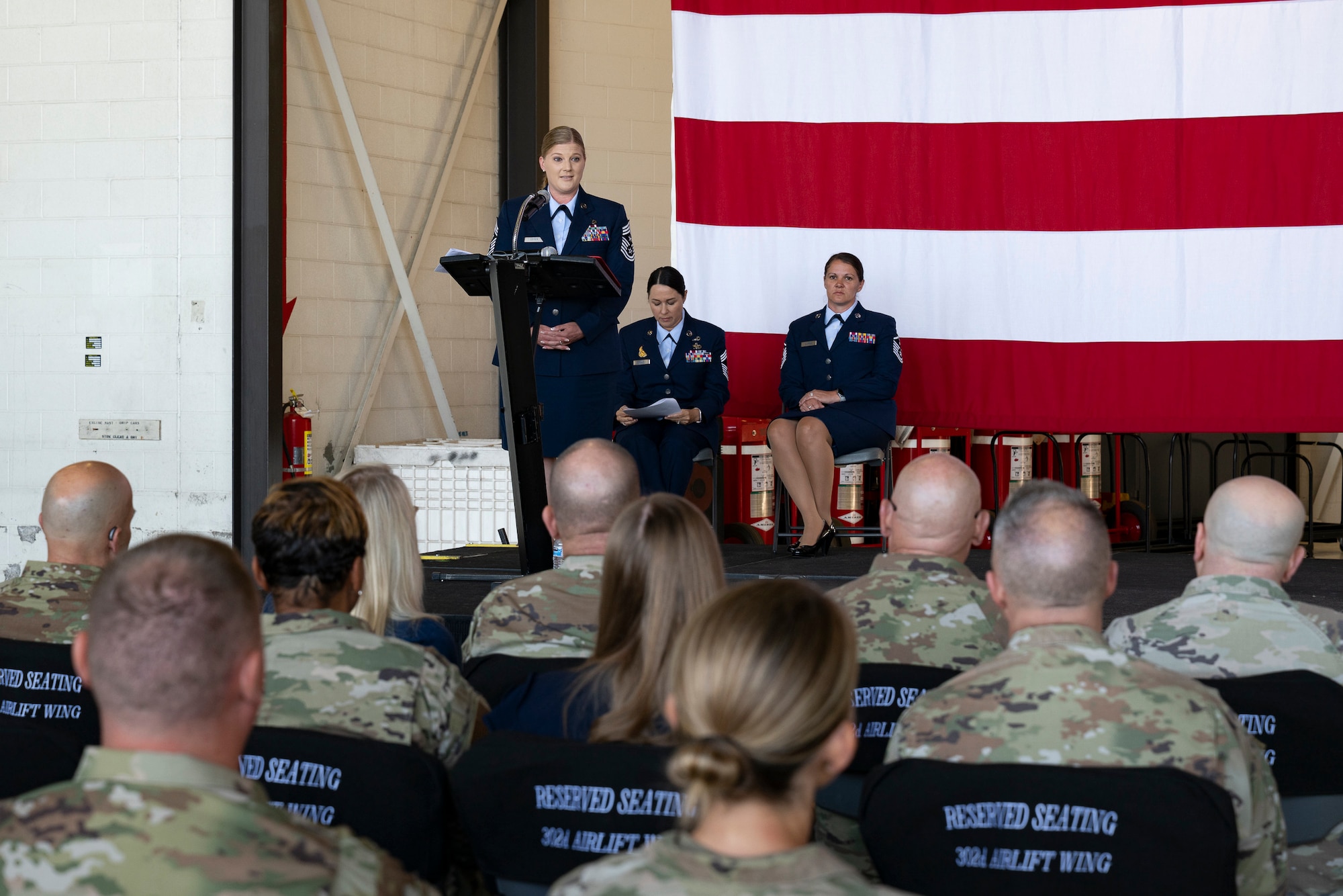 A woman in military service dress standing at a podium on stage with large American flag in the background speaking to a military audience.