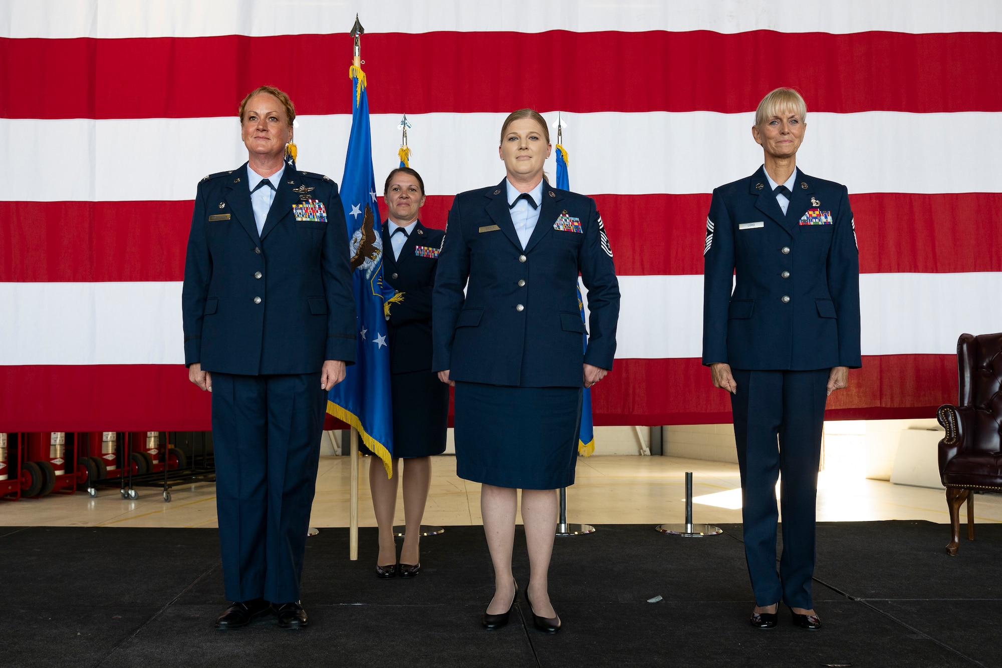Four women in military service dress standing at attention on stage with a large American flag in the background.