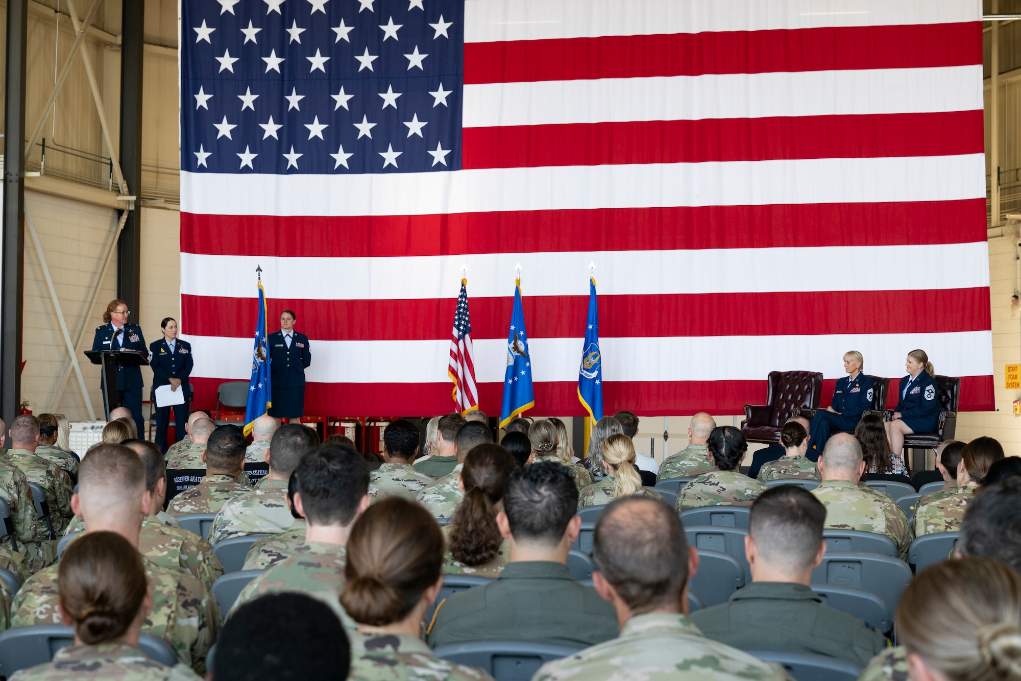 A woman in military service dress on stage speaking to two other women on the other side of the stage with a military audience in attendance and a large American flag in the background.