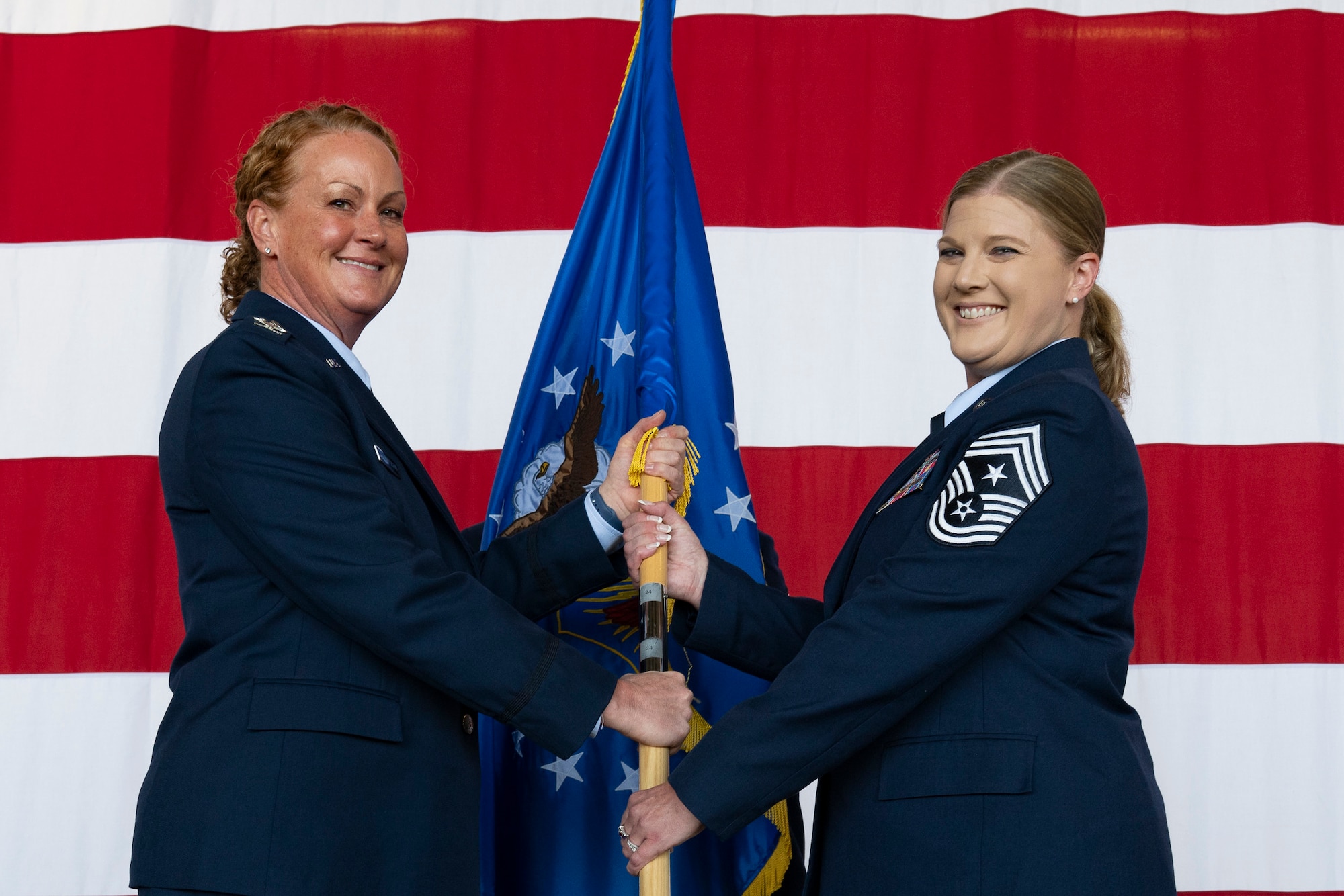 A woman in military service dress handing a flag to another woman in military service dress with a large American flag in the background.