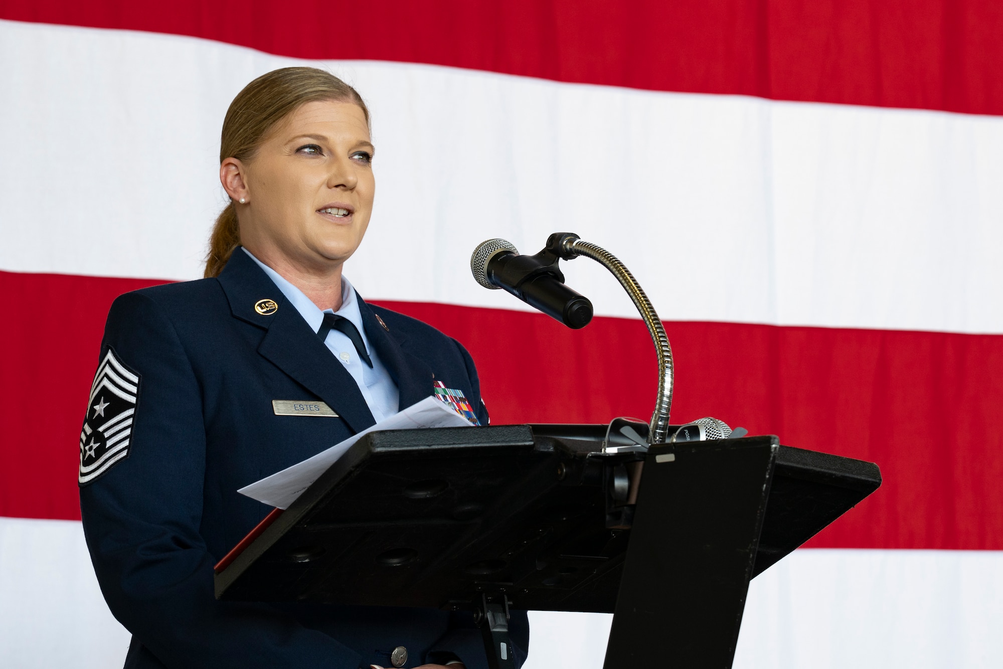 A woman in military service uniform standing in front of a podium with a large American flag behind her.