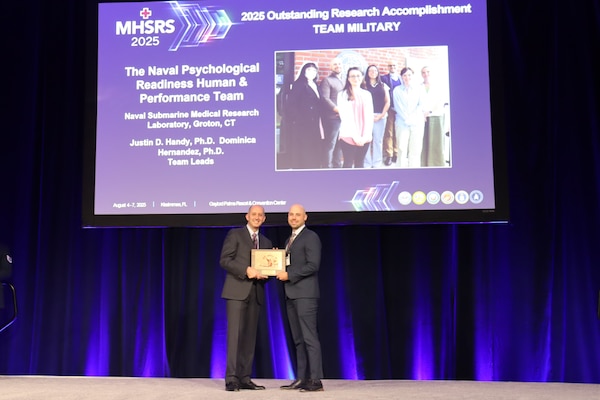 KISSIMMEE, Fla. (Aug. 4, 2025) Dr. Stephen Ferrara, Acting Assistant Secretary of Defense for Health Affairs, presents the Military Health System Research Symposium (MHSRS) award for Outstanding Research Accomplishment by a Military Team to Dr. Justin Handy, principal investigator and representative of the Naval Psychological Readiness and Human Performance (NPRHP) team. This team, from Naval Submarine Medical Research Center (NSMRL), focuses on improving the holistic health of the undersea warfighter, and has received recognition for their work modernizing the Submarine Environment Fit Test (SUBFIT), which was recently implemented throughout the entire Submarine Force (SUBFOR). NSMRL, part of Navy Medicine Research & Development and based out of Groton, Connecticut, sustains the readiness and superiority of U.S. undersea warfighters through innovative health and performance research and works to lead the world in delivering science solutions to ensure undersea warrior dominance. (U.S. Navy Photo by Emily Swedlund/ released)