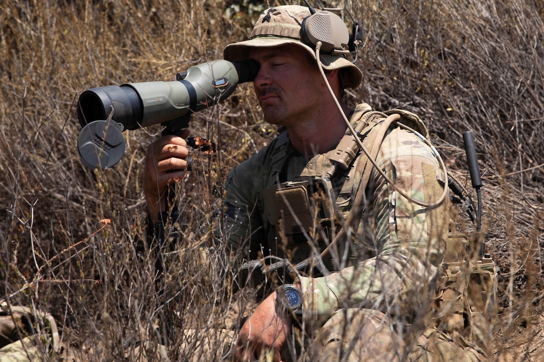 A soldier looks through a magnifying scope using one eye while kneeling outdoors amid branchy vegetation.