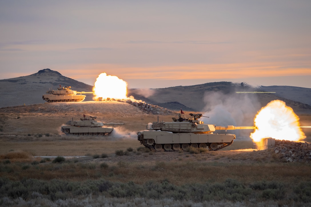 Three tanks fire in the mountains creating orange fireballs under a grayish, pink sky.