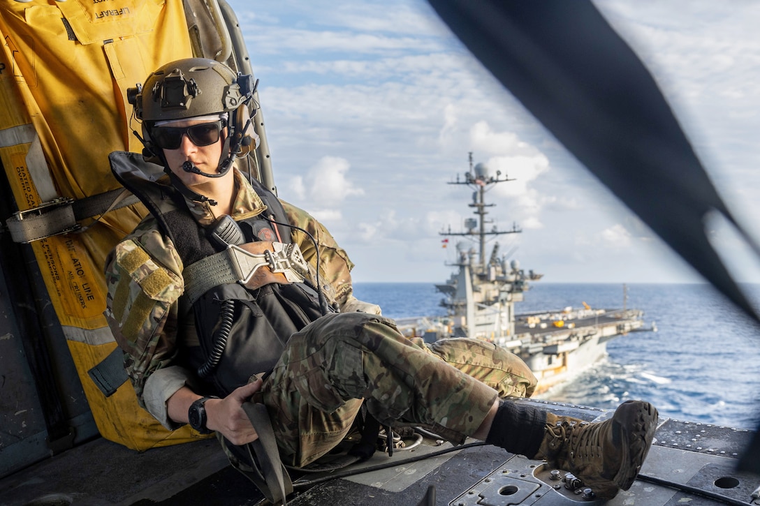 A sailor sits in the open doorway of an airborne helicopter as a partially visible ship transits a body of water in the background.
