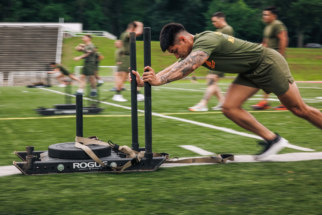 A blurry photo of a Marine pushing a sled on a turf as fellow Marines exercise in the background.