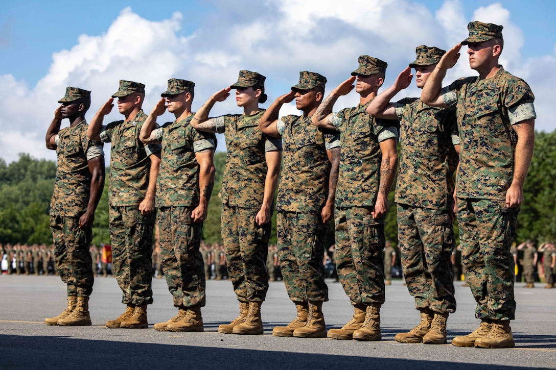 Eight Marines salute while standing in formation on a stage under a blue sky.