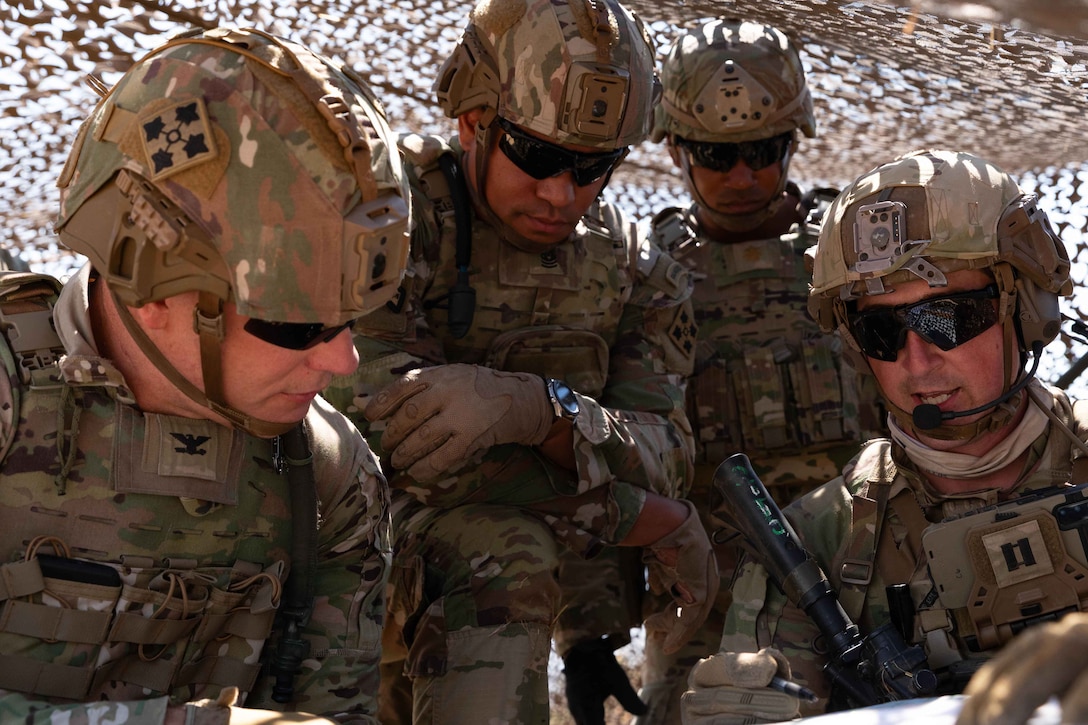 Four soldiers wearing helmets and sunglasses huddle under brown netting and look at an object that one of the soldiers is holding.