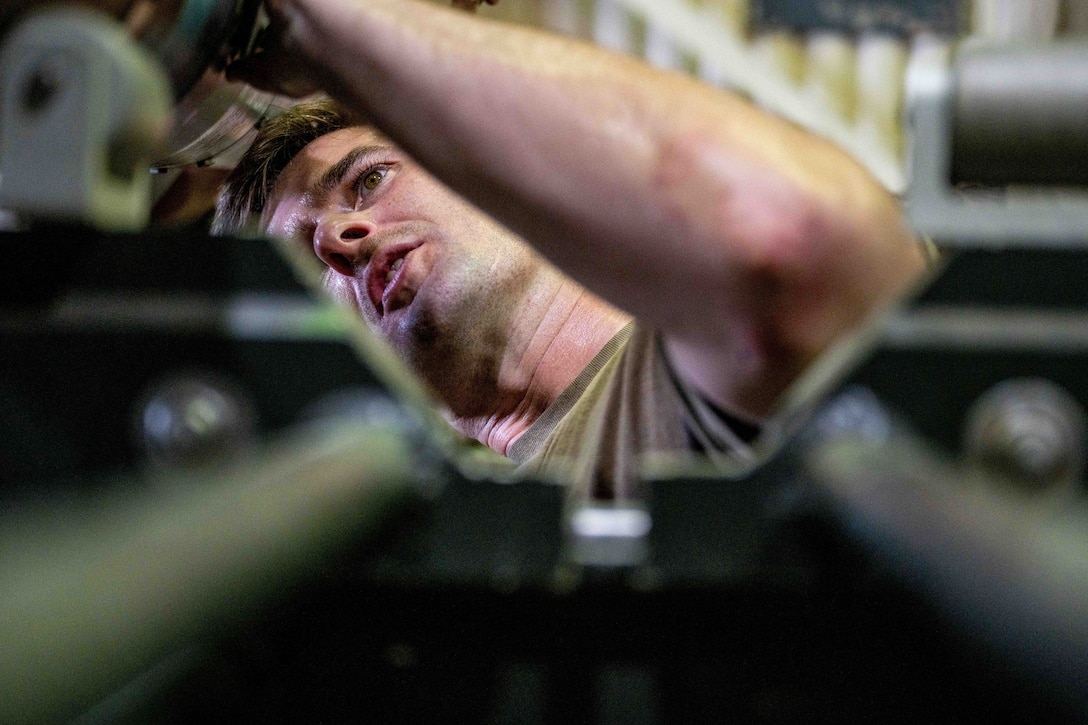 An airman tilts their head and holds up one arm while inspecting a bomb as seen from close up.