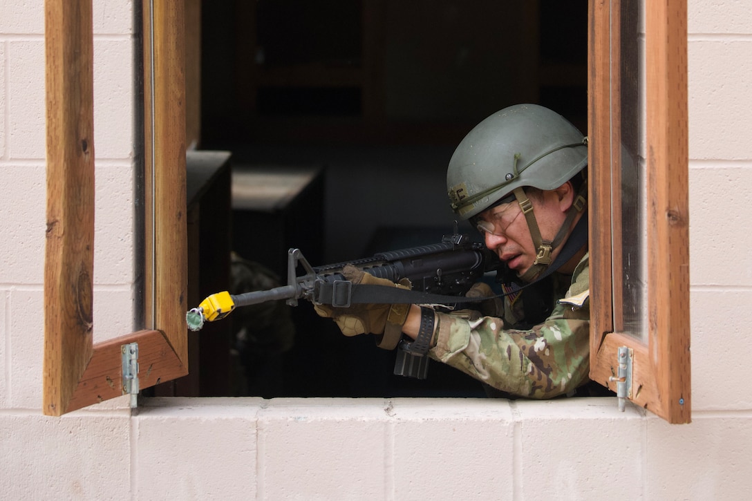 A soldier in a helmet aims a service weapon out of an open window.