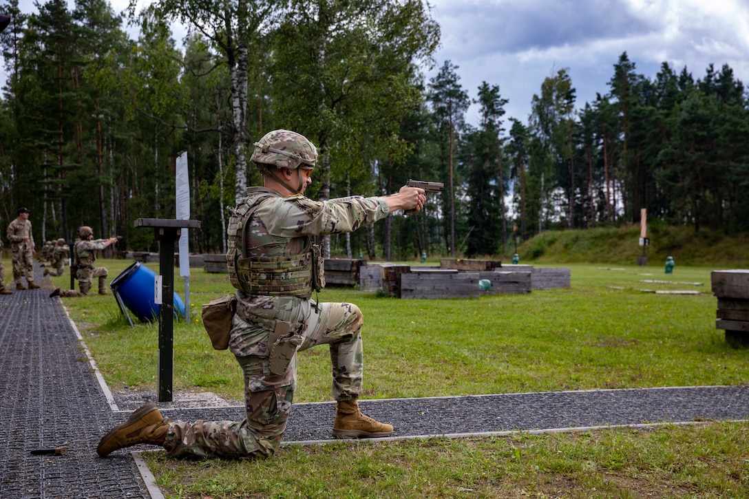 A soldier takes a knee to fire a service weapon in an outdoor course while other soldiers in the background do the same.
