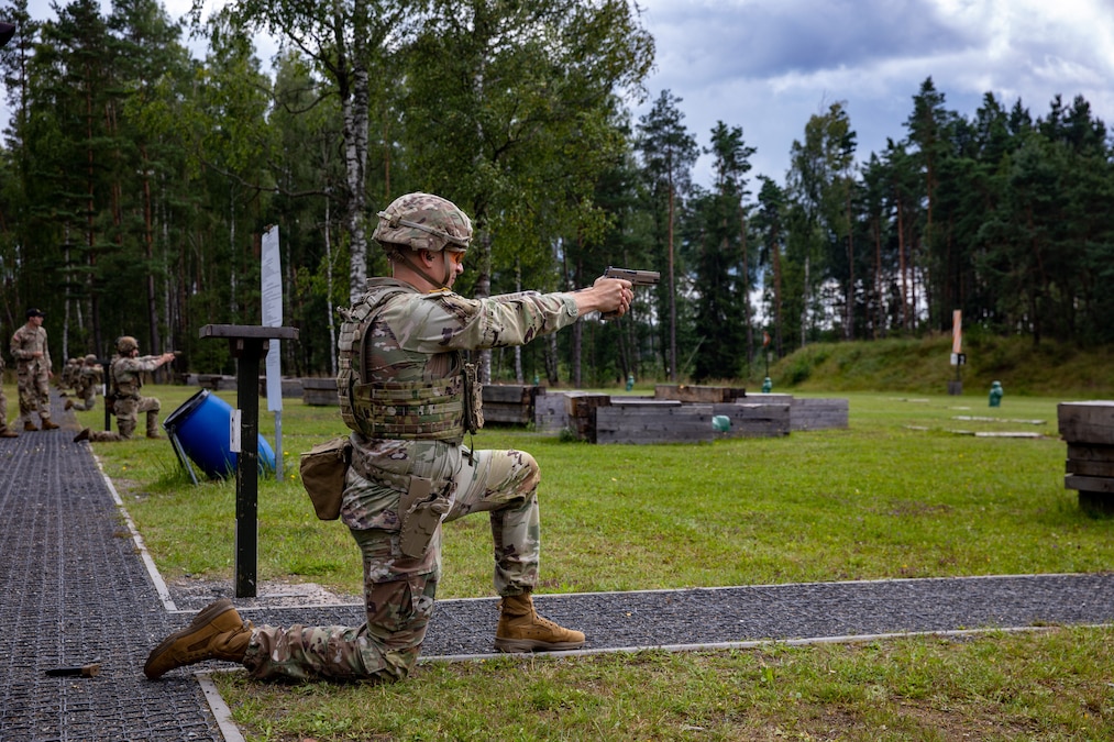 A soldier takes a knee to fire a service weapon in an outdoor course while other soldiers in the background do the same.