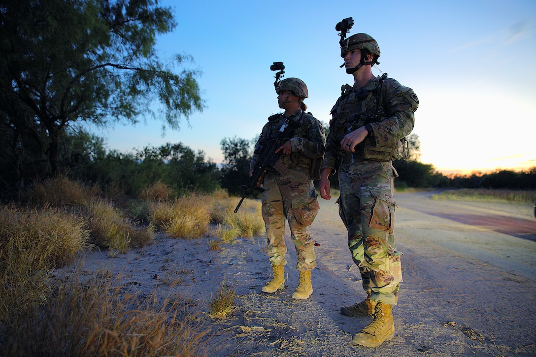 Two soldiers with night-vision goggles attached to their helmets stand on a dirt path with trees and other vegetation in the background as the sun sets behind them.