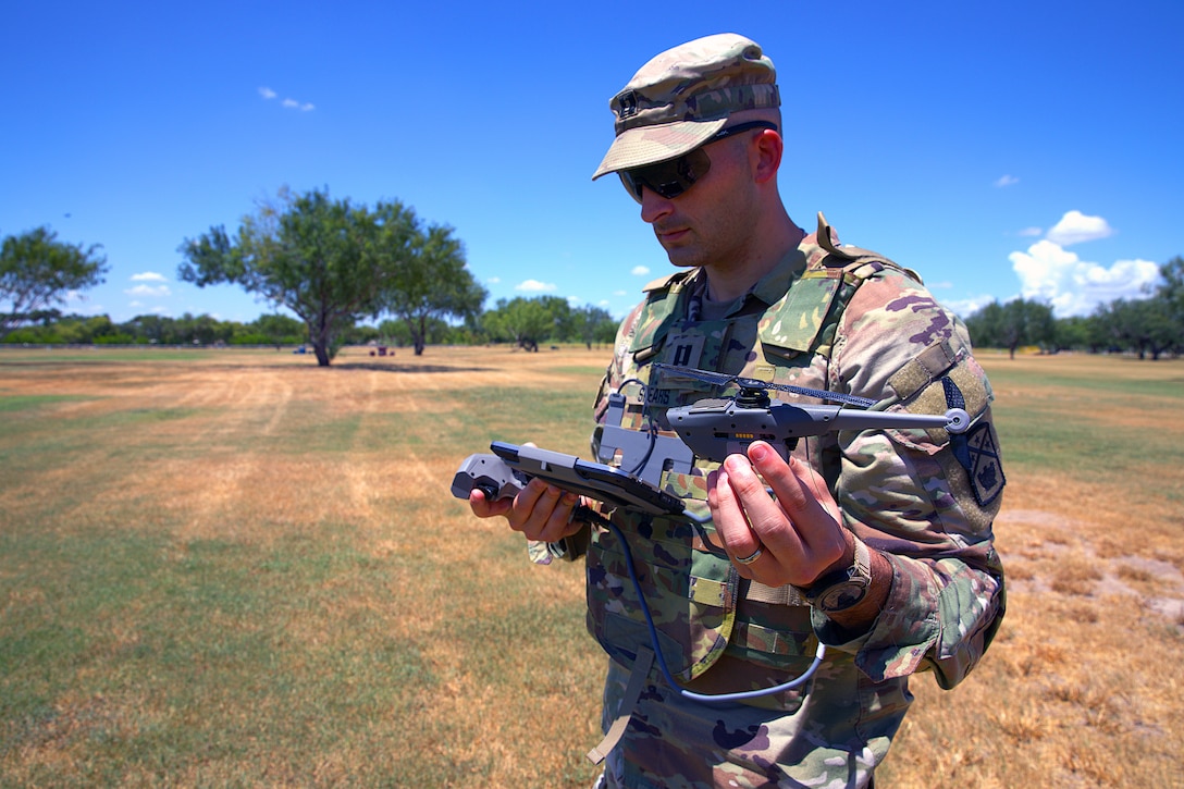 A soldier holds a small drone in one hand and a control device in the other while standing in a grass field on a sunny day; trees are in the background.