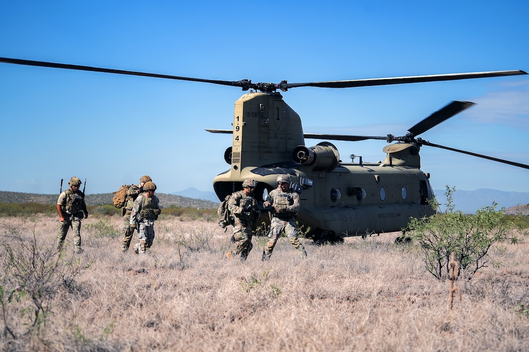 Soldiers and airmen move away from a grounded CH-47 Chinook helicopter in sparsely vegetated terrain, with a blue sky in the background.