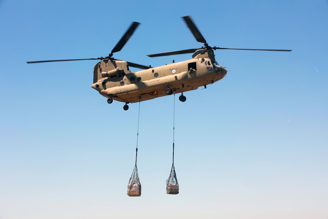 A flying Chinook helicopter carries two containers attached to its underside by cables, with a blue sky in the background.