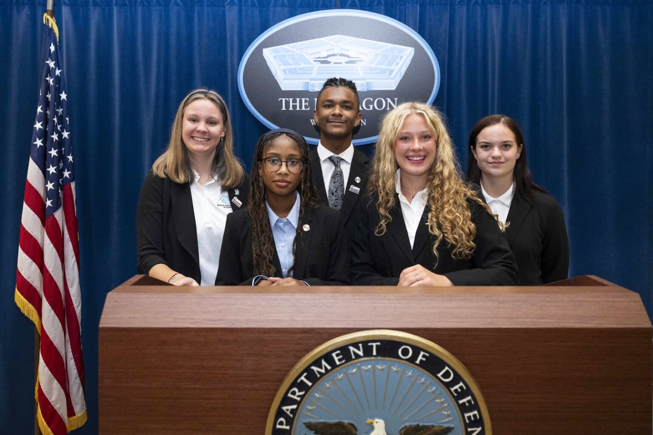 Five young people stand together behind a lectern. Behind them is an American flag and a sign that says, "The Pentagon."