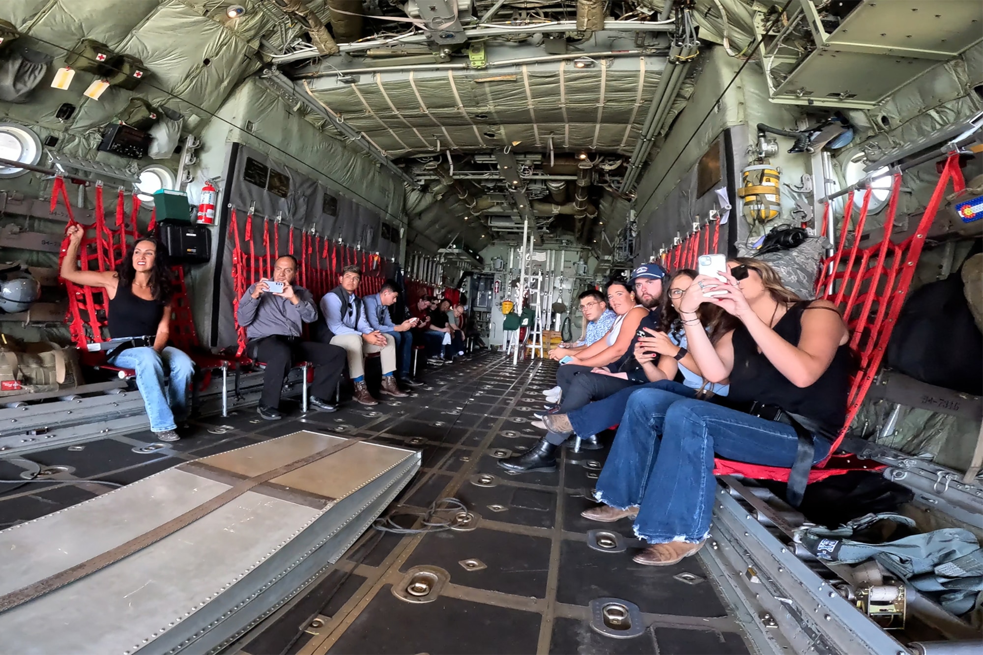 A group of civilians in passenger seating aboard the cargo bay of a military aircraft.