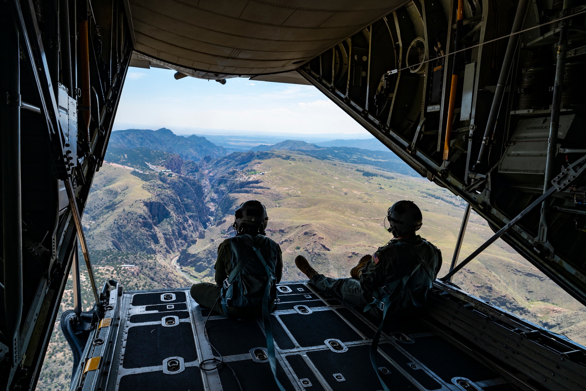 Two people wearing flight suits sitting at the edge of an open aircraft cargo bay ramp door overlooking a mountainous landscape.