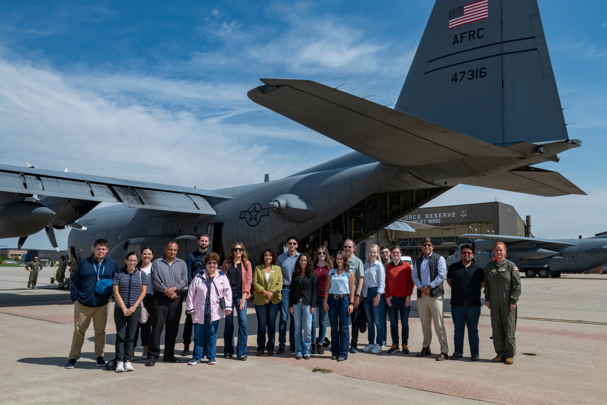 A group of civilians stand next to a woman in a military flight suit next to a military aircraft parked on the flight line.