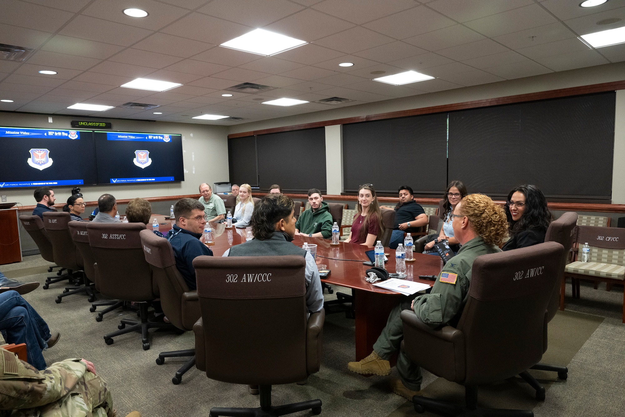 A group of civilians sitting at a large table in a conference room with a woman in a military flight suit at the head of the table.