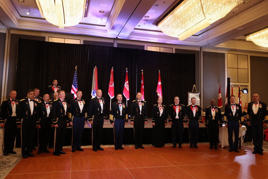 Soldiers and former Soldiers stand in the front of a ball room where there is a stage and flags set up behind them. the Soldiers are in their dress uniforms while the former Soldiers wear suites.