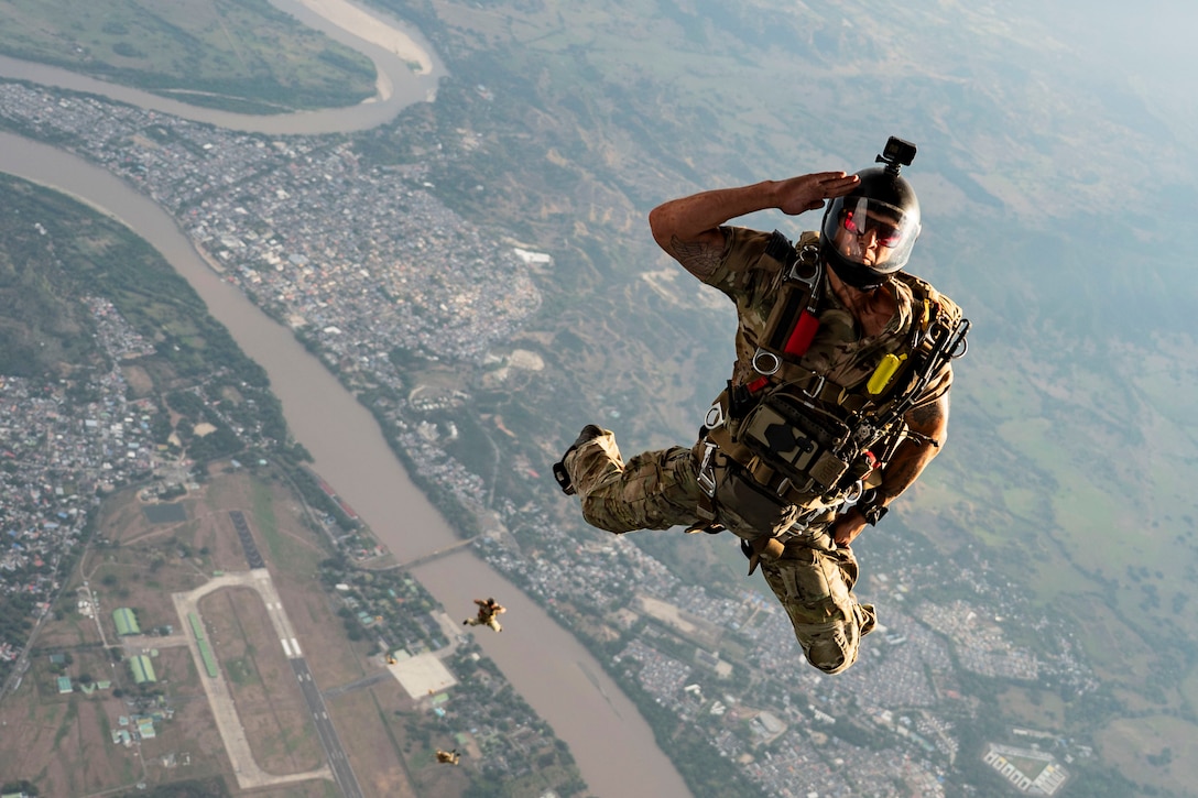 An airman wearing parachuting gear salutes while free falling in a hazy sky over a city.