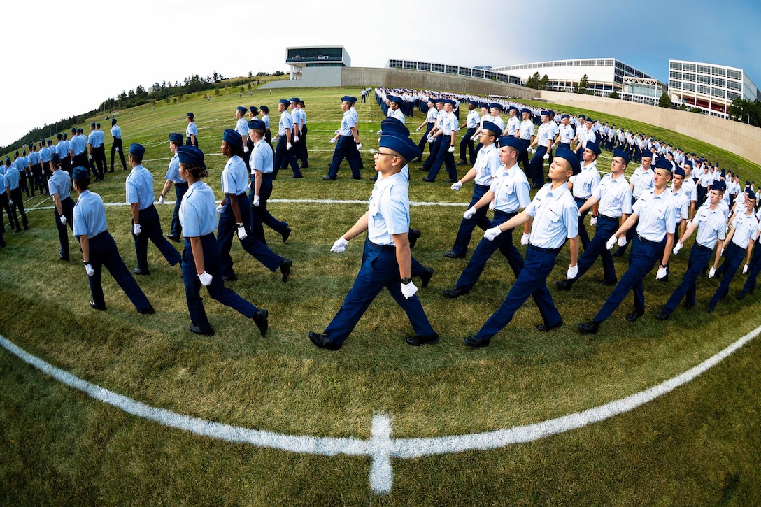A side view of dozens of Air Force Academy cadets march in formation on a turf field outside of a building.