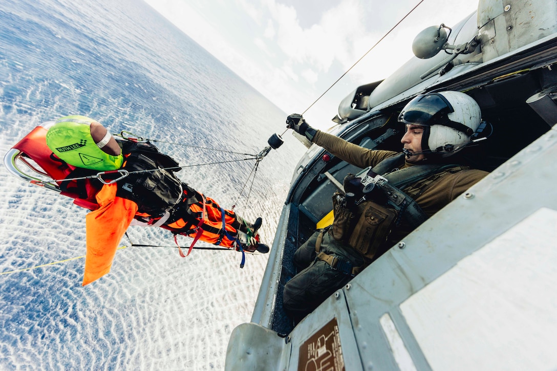 A sailor raises a dummy on a stretcher into an airborne helicopter over a body of water.