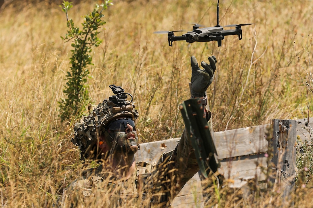 A soldier in camouflage kneels in tall, brown grass while retrieving an airborne drone during the day.