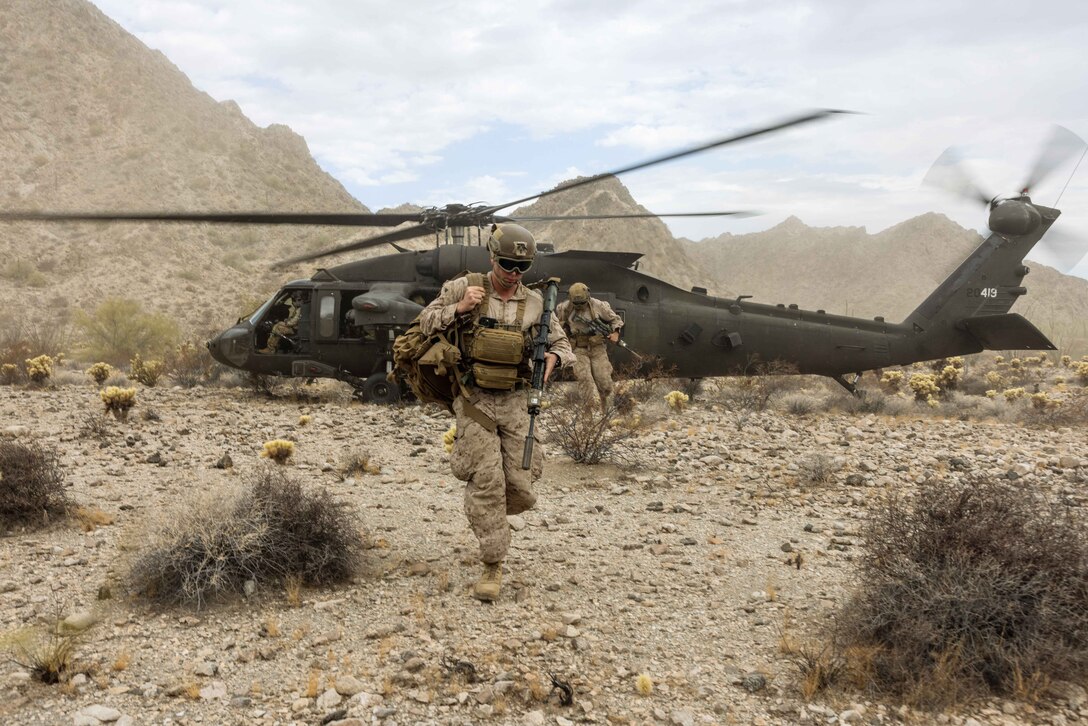 A Marine walks away from a helicopter in a desert-like setting while other Marines follow behind.