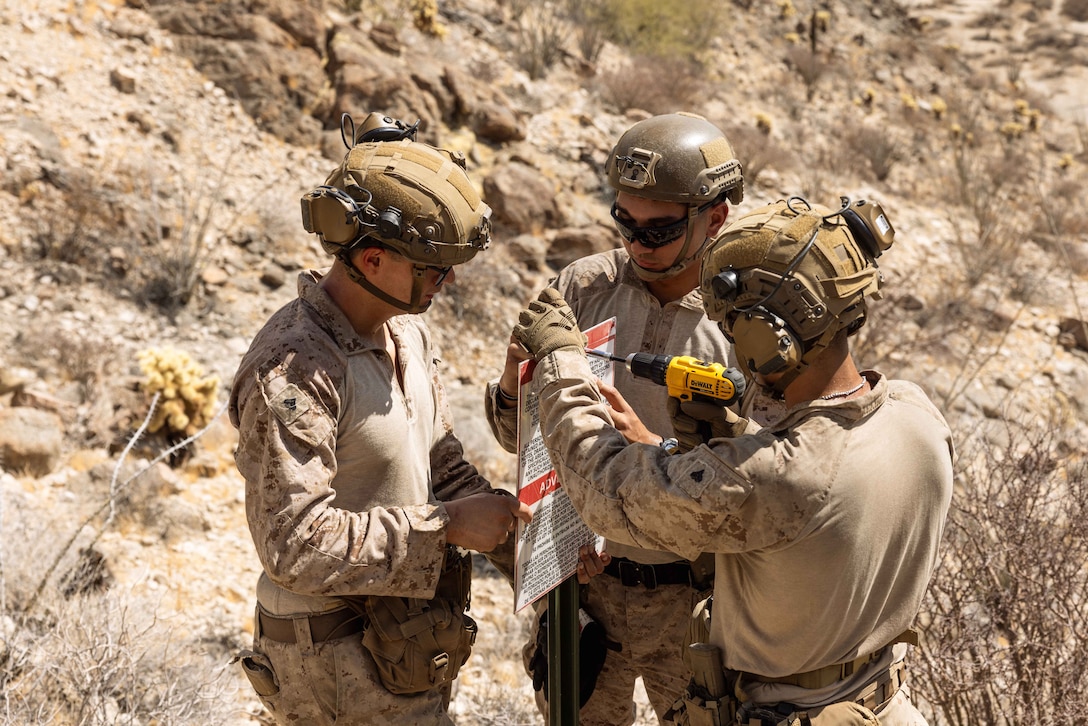Three Marines install a sign on a sunny day in a rocky, desert-like terrain.