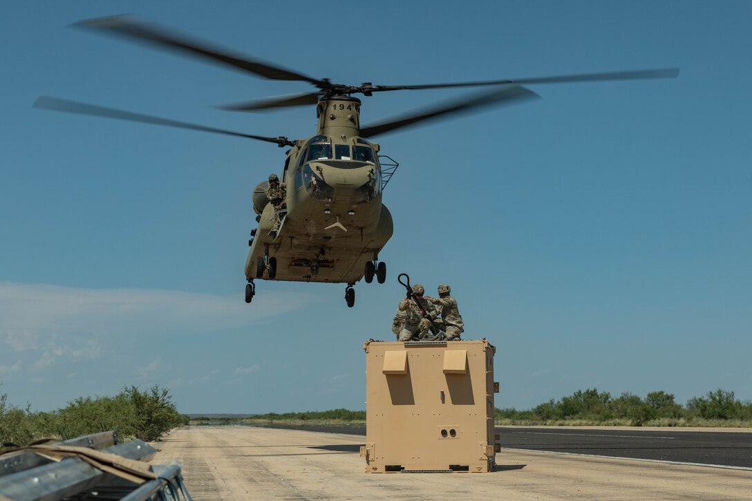 Two soldiers sitting in a metal container reach up towards a helicopter in flight directly above a road on a sunny day.