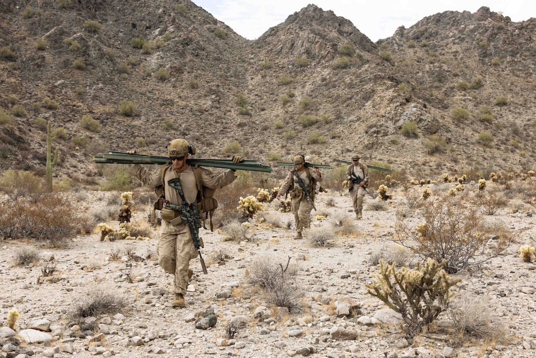 Three Marines walk in a line across rugged terrain holding sign equipment.